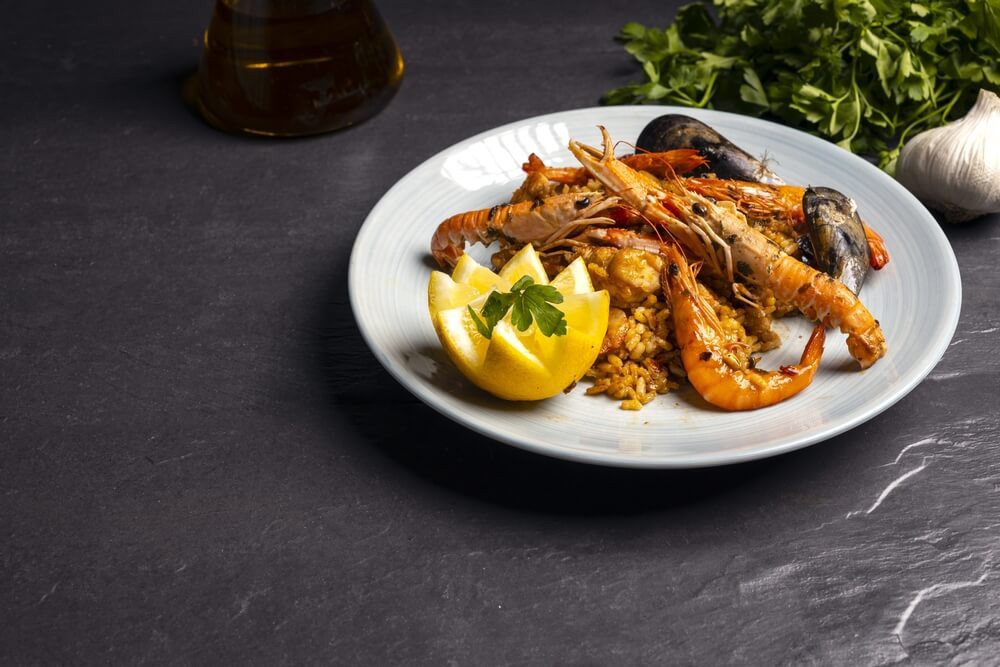 Origins of paella: A plate of paella with king prawns and a lemon on it placed on a black surface