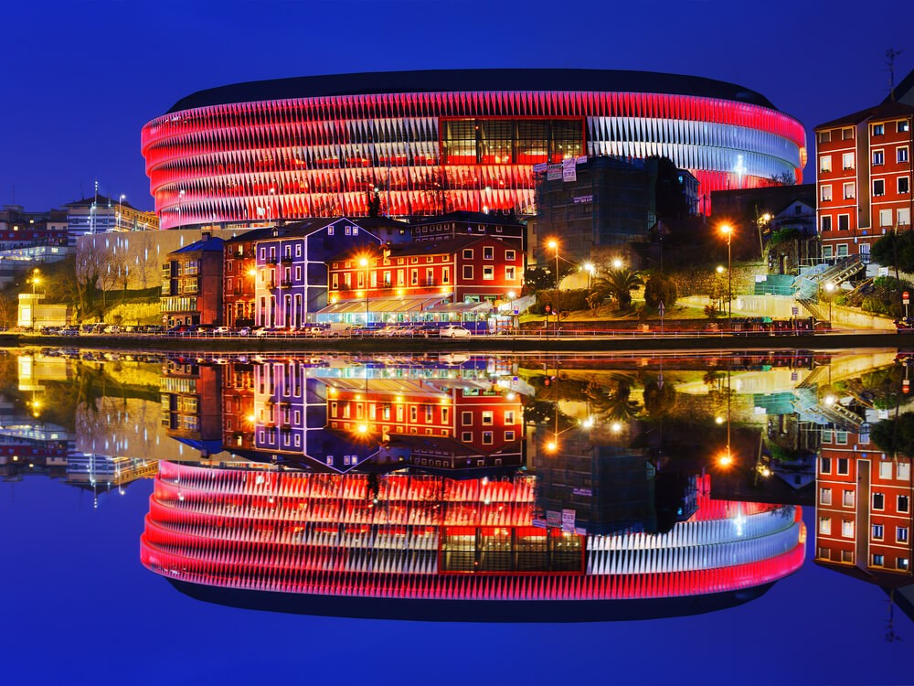 San Mamés Stadium: The curved facade of the football stadium lit up in red and reflected on the river