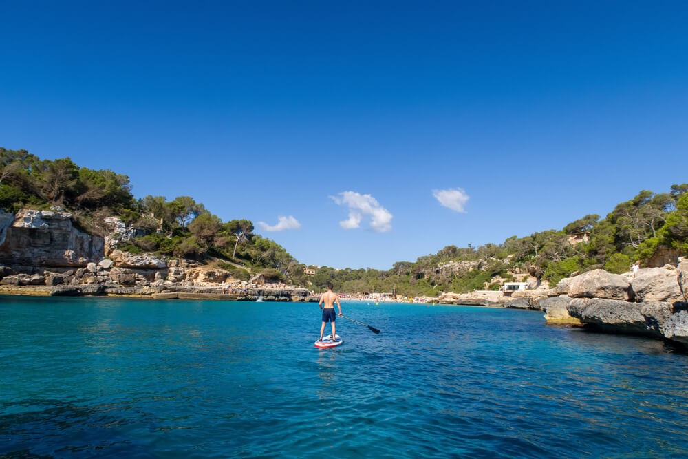 Paddleboarding in Mallorca: Man paddleboarding towards a beach surrounded by rocks