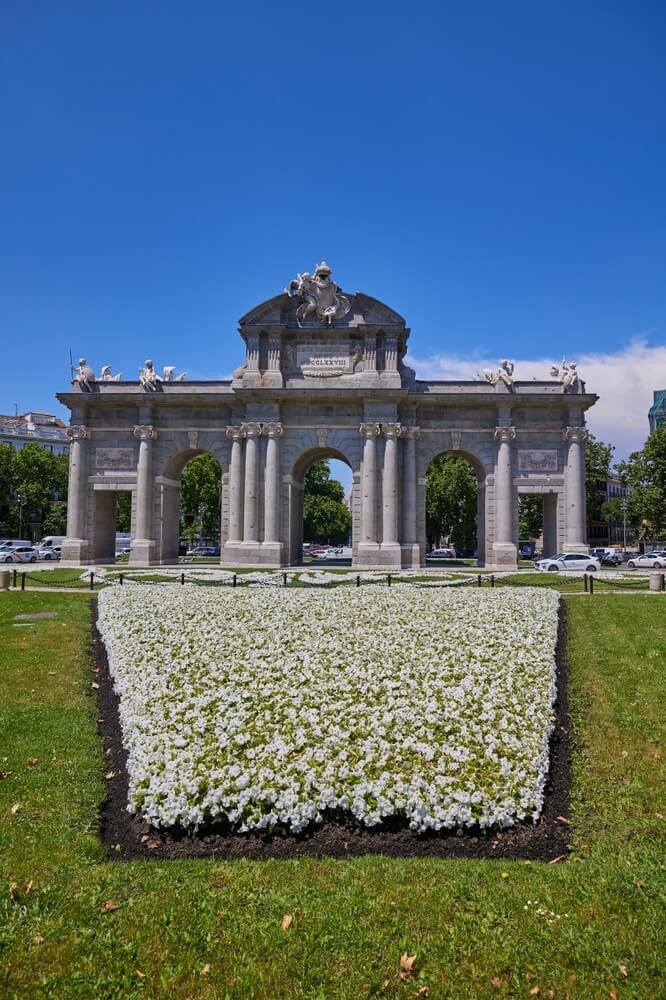 Die Puerta de Alcalá in Madrid mit einem Blumenbeet im Vordergrund.